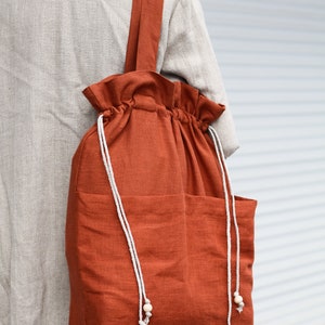 May include: A large, rust-colored canvas tote bag with a drawstring closure and two pockets. The bag has white drawstrings with wooden beads at the ends.