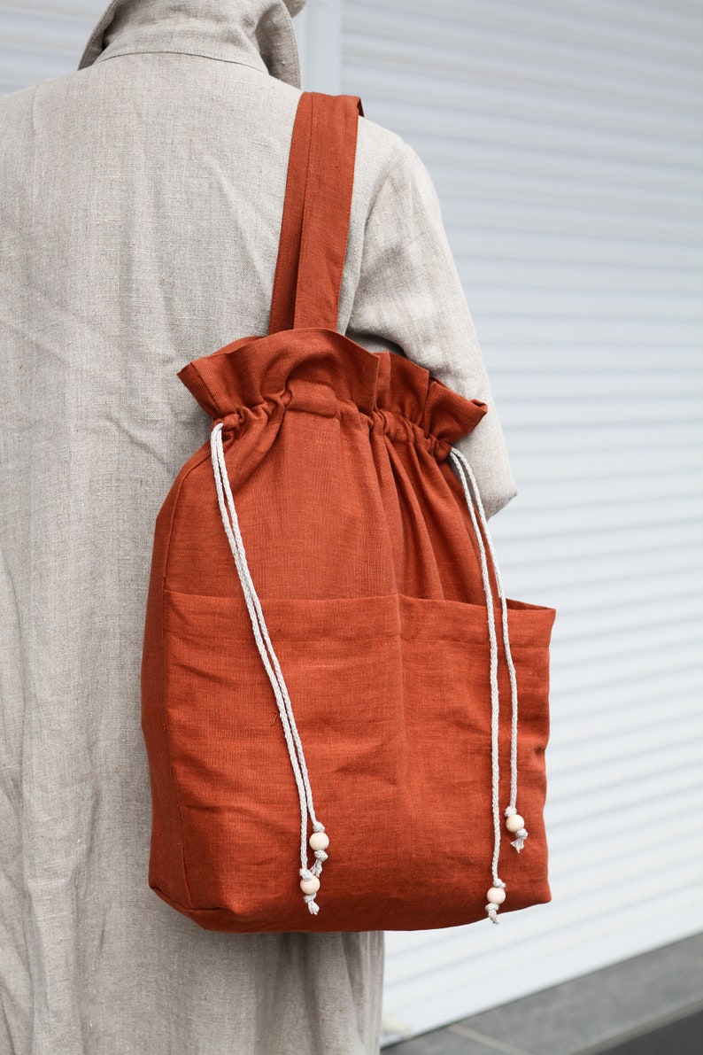 May include: A large, rust-colored canvas tote bag with a drawstring closure and two pockets. The bag has white drawstrings with wooden beads at the ends.