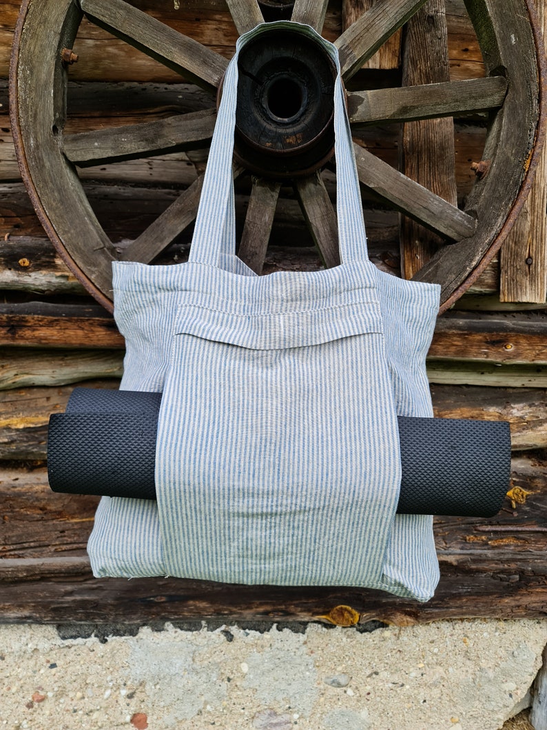 May include: A blue and white striped tote bag with a black yoga mat tucked inside. The bag has a pocket on the front and is hanging from a wooden wheel.