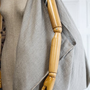 May include: A large, light gray linen tote bag with a wooden arm mannequin inside. The bag has a simple, unstructured design and is made of a natural fabric.