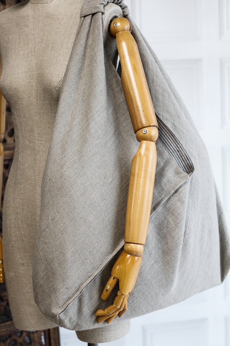 May include: A large, light gray linen tote bag with a wooden arm mannequin inside. The bag has a simple, unstructured design and is made of a natural fabric.