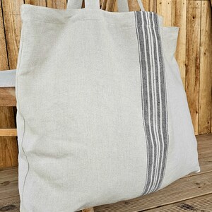 May include: A large, light gray tote bag with long handles. A vertical stripe of dark gray and white stripes runs down the center of the bag. The bag is made of a textured fabric and appears to be empty.