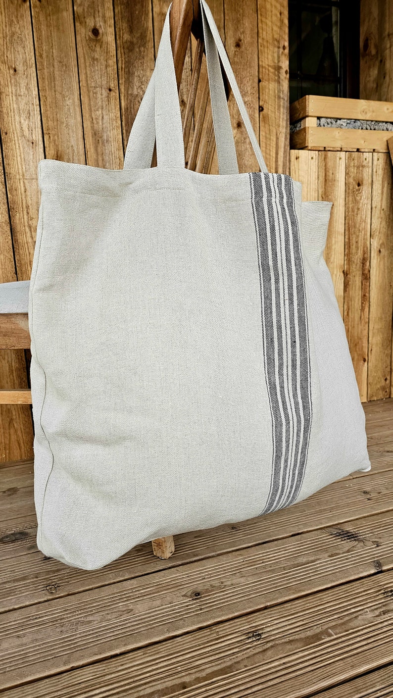 May include: A large, light gray tote bag with long handles. A vertical stripe of dark gray and white stripes runs down the center of the bag. The bag is made of a textured fabric and appears to be empty.