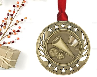 May include: A gold-colored cheerleading medal with a red ribbon. The medal features a megaphone, pom-poms, and flags, surrounded by a laurel wreath and stars. The background includes a white wooden surface, frosted greenery, and berries.