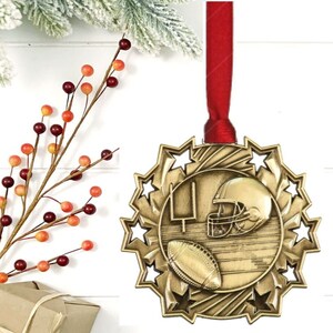 May include: A gold-colored football-themed ornament with a red ribbon for hanging. The ornament features a football, helmet, and goalpost design, surrounded by star cutouts. Perfect for sports fans.