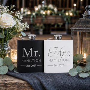 May include: A black flask with the text Mr. HAMILTON Est. 2027 and a white flask with the text Mrs. HAMILTON Est. 2027. Both flasks feature a silver-toned screw-top cap and a rectangular body design, presented as a pair of matching containers.