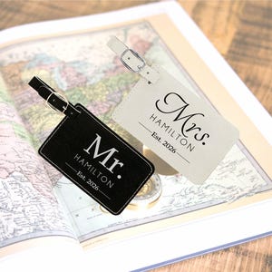 May include: Two personalized luggage tags, one black and one cream, with the text "Mr. Hamilton Est. 2026" and "Mrs. Hamilton Est. 2026". The tags are rectangular with stitched edges and a buckle strap. They rest on an open vintage map.