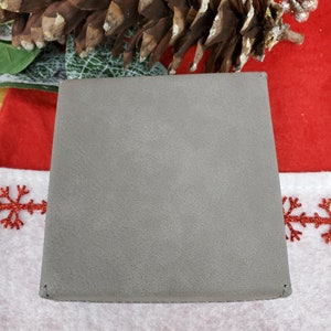 May include: A square, gray wallet with a textured surface. The wallet is closed and appears to be made of a soft material. The background includes a pine cone and festive holiday decorations.