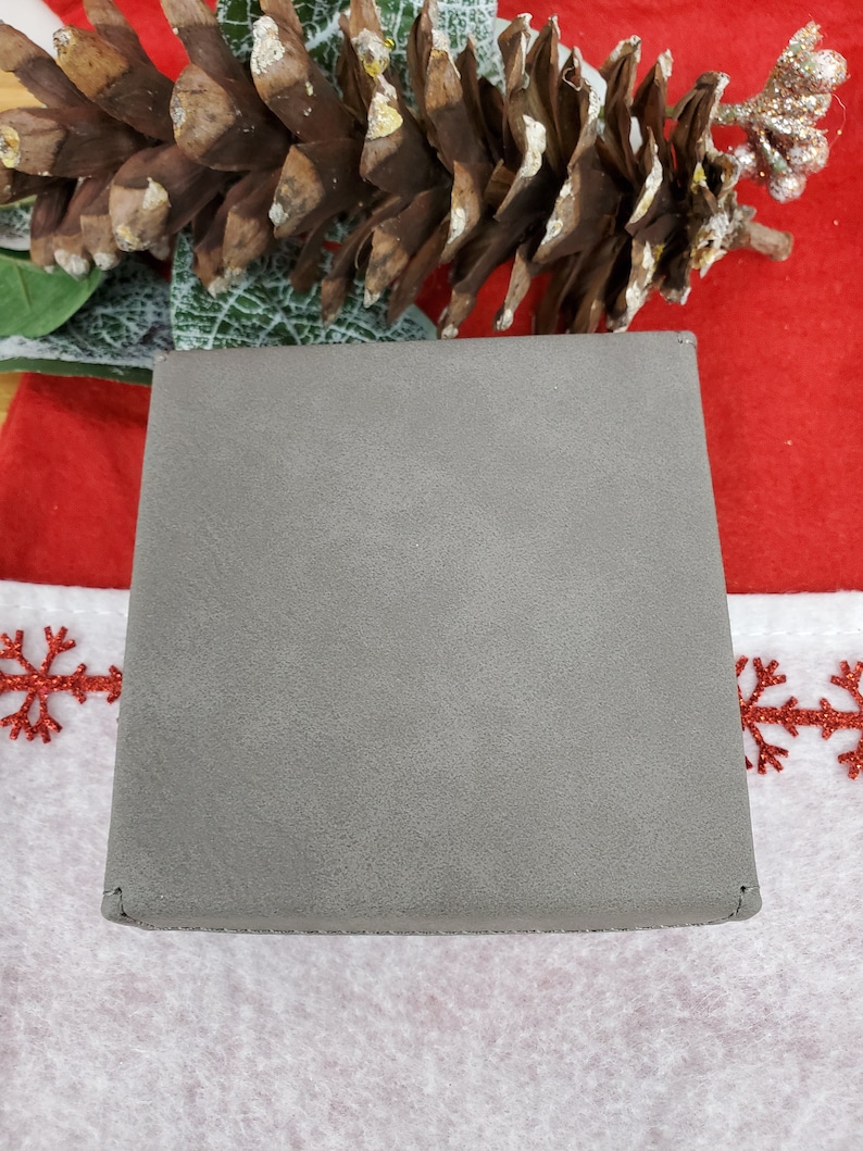 May include: A square, gray wallet with a textured surface. The wallet is closed and appears to be made of a soft material. The background includes a pine cone and festive holiday decorations.