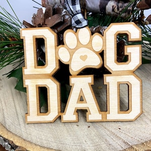 May include: Wooden ornament with the words "Dog Dad" in white letters with a paw print in the center. The paw print has a small heart in the center.