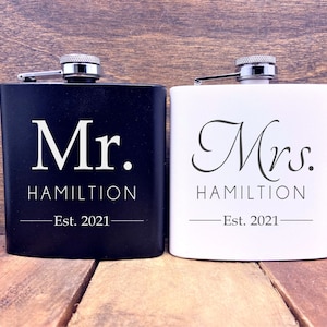 May include: Two black and white hip flasks with silver tops. The black flask has "Mr. HAMILTON Est. 2021" printed on it. The white flask has "Mrs. HAMILTON Est. 2021" printed on it.