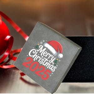 May include: A gray gift box with the text "Merry Christmas 2025" and a Santa hat design. A red Christmas ornament and ribbon are in the background. The box is open, revealing a black interior.