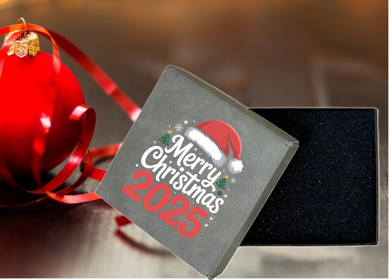 May include: A gray gift box with the text "Merry Christmas 2025" and a Santa hat design. A red Christmas ornament and ribbon are in the background. The box is open, revealing a black interior.