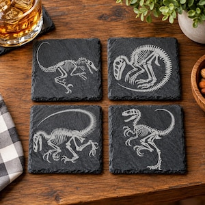 May include: A set of four square black slate coasters, each featuring a distinct white engraved dinosaur skeleton. The designs include a running raptor, a curled raptor, a long-tailed dinosaur in profile, and a standing raptor with claws.