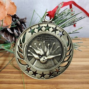 May include: A bronze-colored bowling-themed Christmas ornament. The ornament features a bowling pin design with a ball and pins, surrounded by stars and a laurel wreath. It has a red ribbon for hanging.