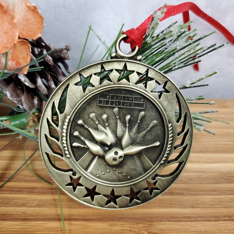 May include: A bronze-colored bowling-themed Christmas ornament. The ornament features a bowling pin design with a ball and pins, surrounded by stars and a laurel wreath. It has a red ribbon for hanging.