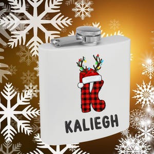 May include: A white flask with a silver cap and hinge. The flask features a Christmas-themed design with a red and black plaid letter "K" wearing a Santa hat and antlers with colorful lights. The name "Kaliegh" is printed below the design.