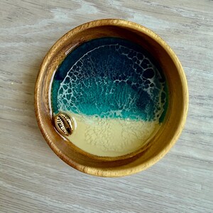 May include: A wooden bowl with a resin design depicting an ocean scene. The bowl features a gold seashell, a sandy beach, and ocean waves in shades of teal and blue. The bowl is round and has a natural wood grain.