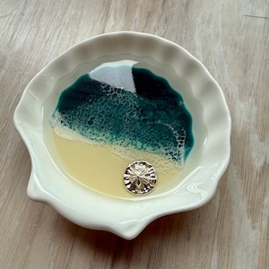 May include: A white, scalloped-edge dish with a beach-themed design. The interior features a yellow 'sand' area, a teal and white 'ocean' section, and a gold-colored sand dollar. The dish is likely used for decorative purposes.