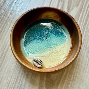 May include: A wooden bowl with a resin design depicting a beach scene. The bowl's interior features a gradient of blue and yellow, with a silver seashell. The bowl is made of wood.