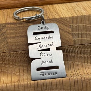 May include: A silver-colored metal keychain with a ring at the top. The keychain is engraved with the names Emily, Samantha, Michael, Olivia, Jacob, and Brianna. The keychain is displayed on a wooden surface.