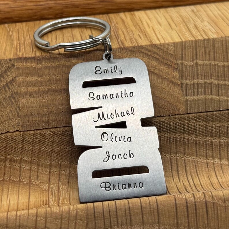 May include: A silver-colored metal keychain with a ring at the top. The keychain is engraved with the names Emily, Samantha, Michael, Olivia, Jacob, and Brianna. The keychain is displayed on a wooden surface.