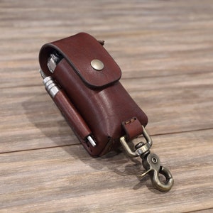May include: A brown leather multi-tool pouch with a metal snap hook. The pouch has a flap with a snap closure and a small pocket on the front. A silver multi-tool and a silver pen are visible inside the pouch.