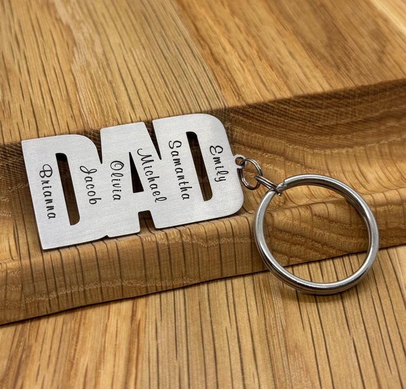 May include: A silver-colored keychain in the shape of the word "DAD" with the names Brianna, Jacob, Olivia, Michael, Samantha, and Emily engraved on it. The keychain is attached to a silver key ring. The background is a wooden surface.