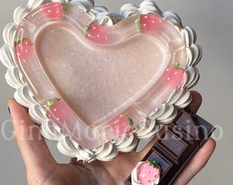 May include: A heart-shaped ashtray with a translucent pink base, embedded with small pink strawberry graphics, and bordered by white piped frosting. A dark brown rectangular chocolate bar accessory features a single strawberry and white frosting.