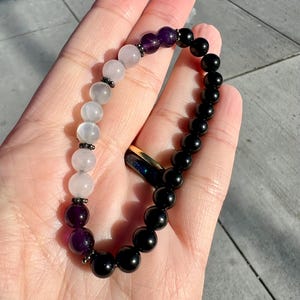 Puede incluir: Un brazalete de cuentas con cuentas negras, blancas y moradas. El brazalete está hecho de piedras naturales.
