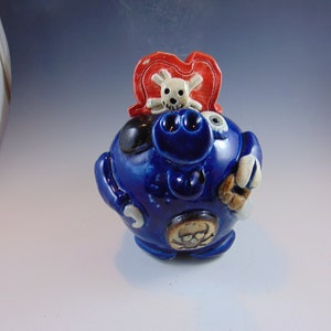 May include: Blue ceramic piggy bank with a skull and crossbones design on the top. The piggy bank has a white and red hat on top of the skull.