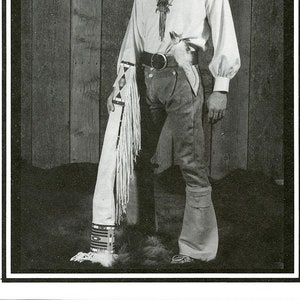 May include: A man wearing a white shirt with a beaded necklace and a fringed leather vest. He is holding a white fringed bag in his left hand. The background is a wooden wall.