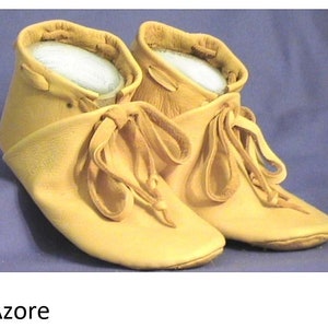 May include: A pair of tan leather moccasins with laces tied in a bow. The moccasins have a soft, suede-like texture and are made of a single piece of leather.