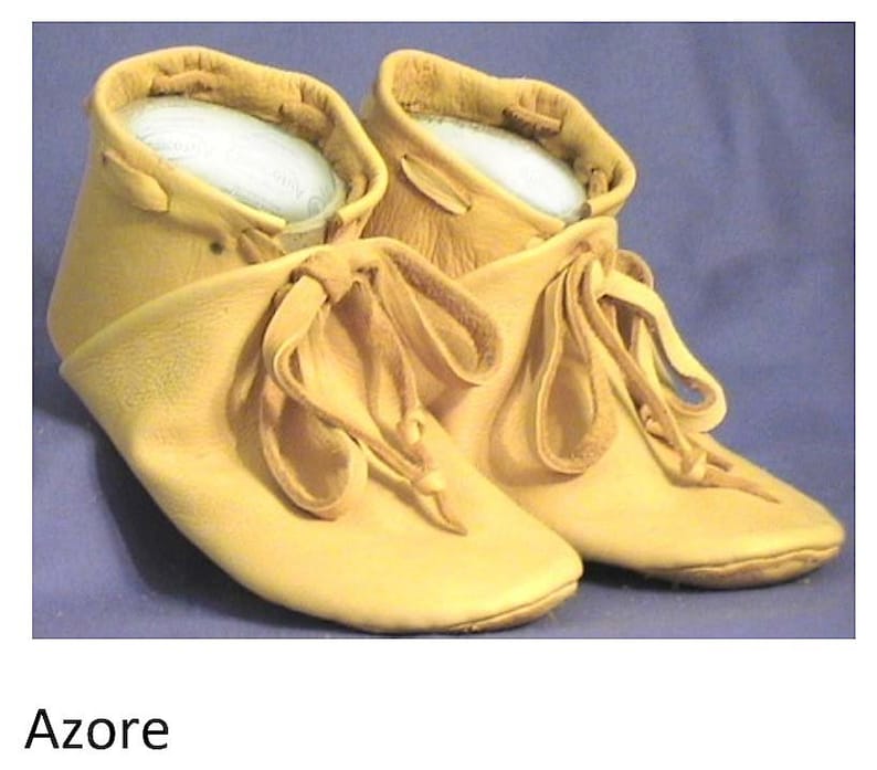 May include: A pair of tan leather moccasins with laces tied in a bow. The moccasins have a soft, suede-like texture and are made of a single piece of leather.