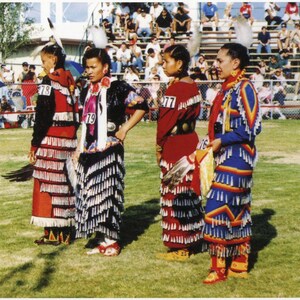 May include: Four young people in traditional Native American regalia, wearing colorful beaded dresses with fringe, stand on a grassy field. The dresses are adorned with feathers and other decorative elements. The people are smiling and appear to be enjoying themselves.