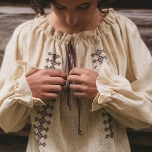 Pode incluir: Uma blusa de linho cor creme com padr&otilde;es geom&eacute;tricos bordados em marrom e preto. A blusa tem um decote franzido e mangas compridas com babados nos punhos.