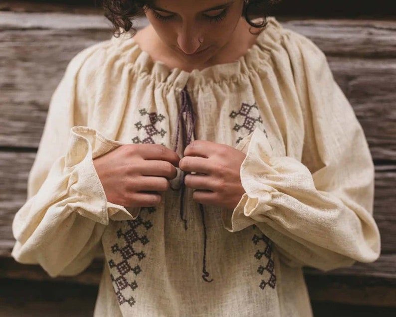 Pode incluir: Uma blusa de linho cor creme com padr&otilde;es geom&eacute;tricos bordados em marrom e preto. A blusa tem um decote franzido e mangas compridas com babados nos punhos.
