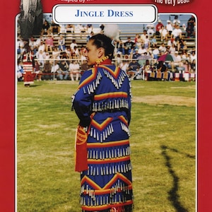 May include: A blue and red jingle dress with silver bells and yellow trim. The dress is worn by a person standing on a grassy field. The text "Jingle Dress" is printed on a white banner above the person.