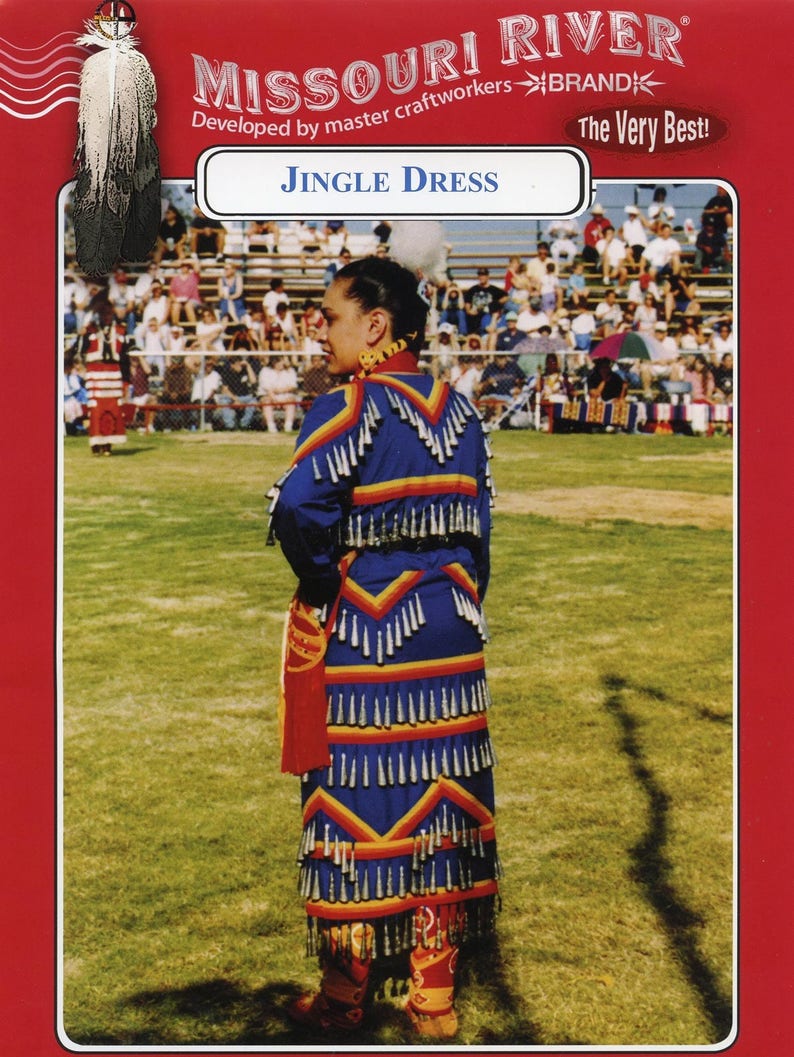 May include: A blue and red jingle dress with silver bells and yellow trim. The dress is worn by a person standing on a grassy field. The text "Jingle Dress" is printed on a white banner above the person.