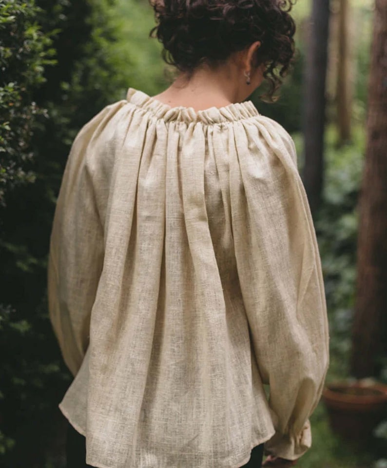 Pode incluir: Uma blusa de linho cor creme com decote franzido e mangas compridas. A blusa &eacute; feita de um tecido leve e respir&aacute;vel e tem um caimento solto.