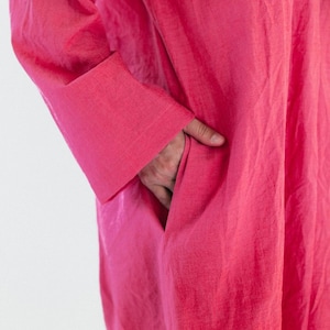 May include: A vibrant fuchsia-colored dress with long sleeves and a pocket. The fabric appears to be lightweight and flowing. The sleeve cuff is visible, and a hand is in the pocket.