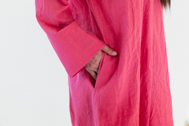 May include: A vibrant fuchsia-colored dress with long sleeves and a pocket. The fabric appears to be lightweight and flowing. The sleeve cuff is visible, and a hand is in the pocket.