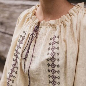 Pode incluir: Uma blusa de linho branca com decote franzido e padr&otilde;es geom&eacute;tricos bordados em tons de roxo e verde.