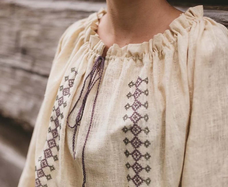Pode incluir: Uma blusa de linho branca com decote franzido e padr&otilde;es geom&eacute;tricos bordados em tons de roxo e verde.