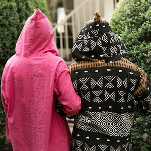 May include: Two hooded robes. One is a vibrant pink, the other is black with a white geometric pattern. The robes are worn by two people standing close together, their backs to the camera. The scene is outdoors, with green foliage in the background.
