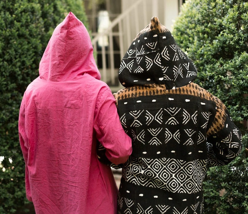 May include: Two hooded robes. One is a vibrant pink, the other is black with a white geometric pattern. The robes are worn by two people standing close together, their backs to the camera. The scene is outdoors, with green foliage in the background.