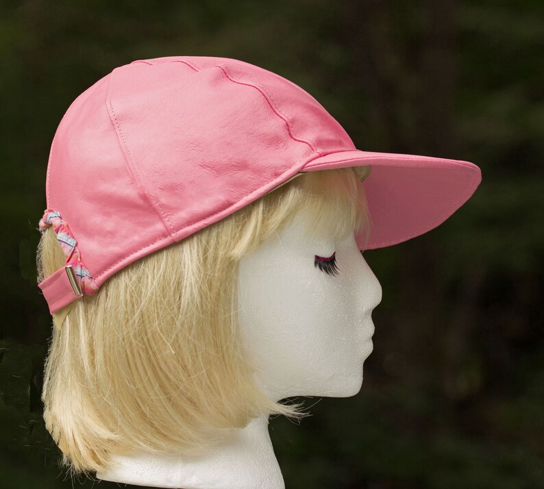 May include: A pink leather baseball cap with a white and blue braided strap. The cap is on a mannequin head with blonde hair.