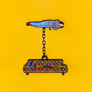 May include: A black enamel pin featuring a couch with a patterned fabric and a person hanging from the ceiling by a chain. The person is lying on their back with their arms crossed.