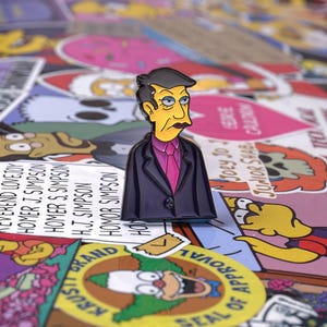 Puede incluir: Un pin de esmalte azul con un personaje de dibujos animados con cabello negro, un traje azul y una corbata rosa. El personaje tiene una expresión seria y mira hacia la derecha. El pin está sobre un fondo de pegatinas de colores.