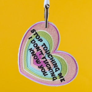 May include: Embroidered heart-shaped patch in pastel colors, featuring the text "STOP TOUCHING ME I DON'T FUCKING KNOW YOU" in black. The patch is held by a metal clip with a white handle against a yellow background.